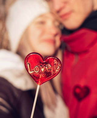 Blurred young couple in love on a date on a winter day.In the foreground is a red lollipop in the shape of a heart and the inscription Love is.Valentine's day concept.