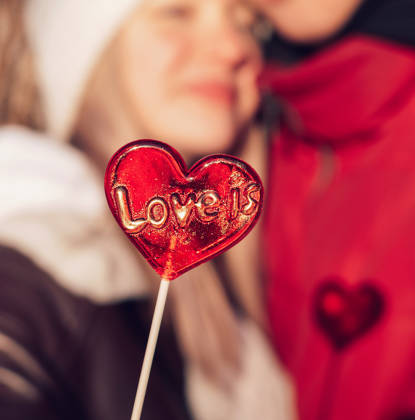 Blurred young couple in love on a date on a winter day.In the foreground is a red lollipop in the shape of a heart and the inscription Love is.Valentine's day concept.