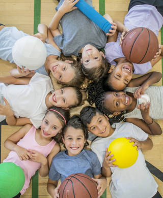 A multi-ethnic group of children are indoors in a health center. They are wearing casual clothing and running shoes. They are lying on the floor with various sports balls, and smiling up at the camera.
