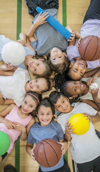 A multi-ethnic group of children are indoors in a health center. They are wearing casual clothing and running shoes. They are lying on the floor with various sports balls, and smiling up at the camera.
