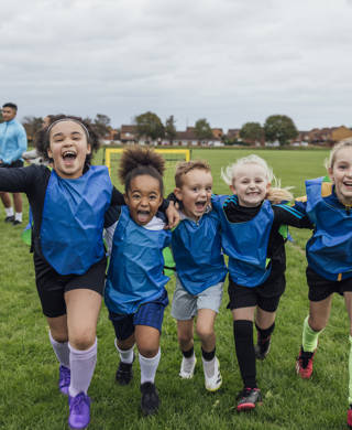 Front view of a small group of boys and girls wearing sports clothing, football boots and a sports bib on a football pitch in the North East of England. They are at football training where they are doing different football training drills. They are running and celebrating with their arms around each other in front of a football goal while smiling and looking at the camera.
Videos are available for this scenario.