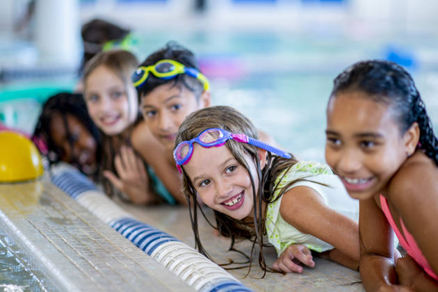 A multi-ethnic group of kids are indoors in a pool. Some of them are wearing goggles and smiling at the camera.