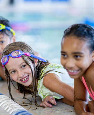 A multi-ethnic group of kids are indoors in a pool. Some of them are wearing goggles and smiling at the camera.