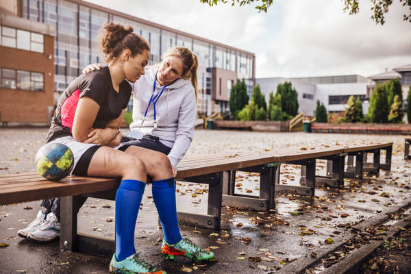 Teenage girl sitting on a bench with her rugby ball at one side. Dirty with mud after her match. Female coach sits close supporting her as the girl looks sad.