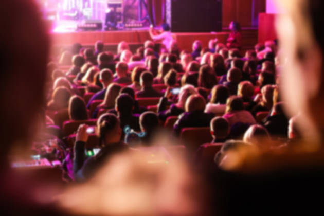a large audience crowd seated in a majestic theater, theatre, auditorium. the people blurred to highlight the stage or screen for a show, concert, or film festival.