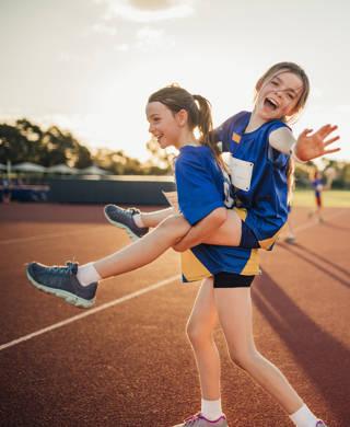 Two girls are messing around at athletics club on the running track. One is giving the other a piggy back and she is reaching out for the camera.