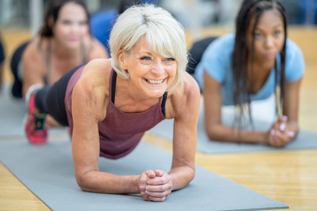 A fit and athletic senior Caucasian woman is participating in a multi ethnic group fitness class at a gym. She is holding a plank position on her yoga mat. She is smiling and happy.