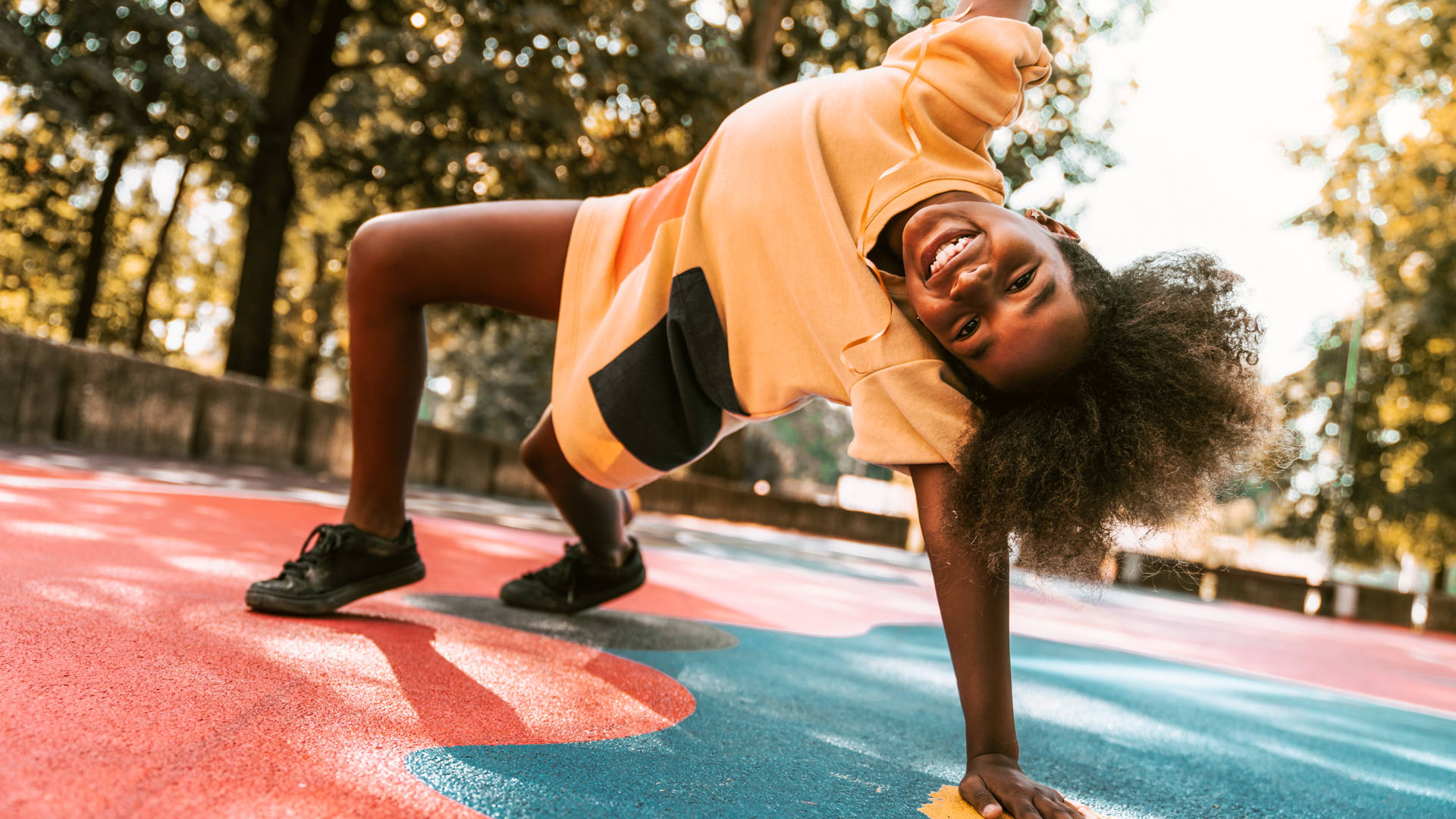 A young girl is playing around the basketball court. She is stretching on one arm, the other is high in the air. She is happy to be outside on a warm summer day.