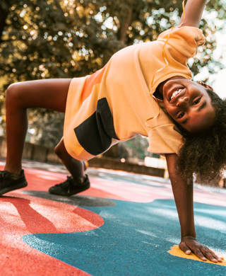 A young girl is playing around the basketball court. She is stretching on one arm, the other is high in the air. She is happy to be outside on a warm summer day.