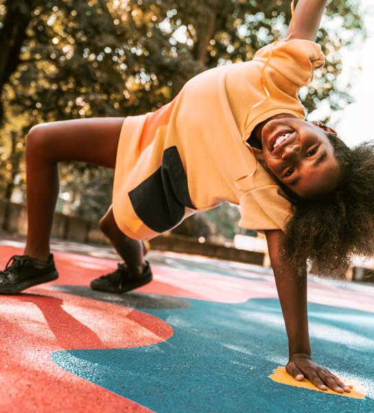 A young girl is playing around the basketball court. She is stretching on one arm, the other is high in the air. She is happy to be outside on a warm summer day.