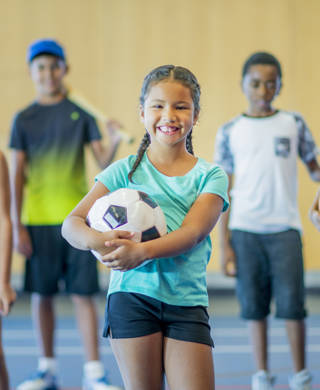 A multi-ethnic group of kids are standing in a gymnasium. They are holding a basketball, volleyball, hockey stick, skipping rope, baseball bat, and soccer ball. They are smiling.