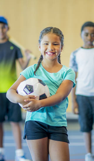 A multi-ethnic group of kids are standing in a gymnasium. They are holding a basketball, volleyball, hockey stick, skipping rope, baseball bat, and soccer ball. They are smiling.