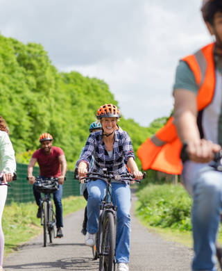 A head-on shot of a small group of people cycling along a path whilst on a community bicycle ride in Hexham, North East England. They are smiling as they cycle, wearing casual clothing and bicycle helmets. The male at the front is wearing a reflective waistcoat.