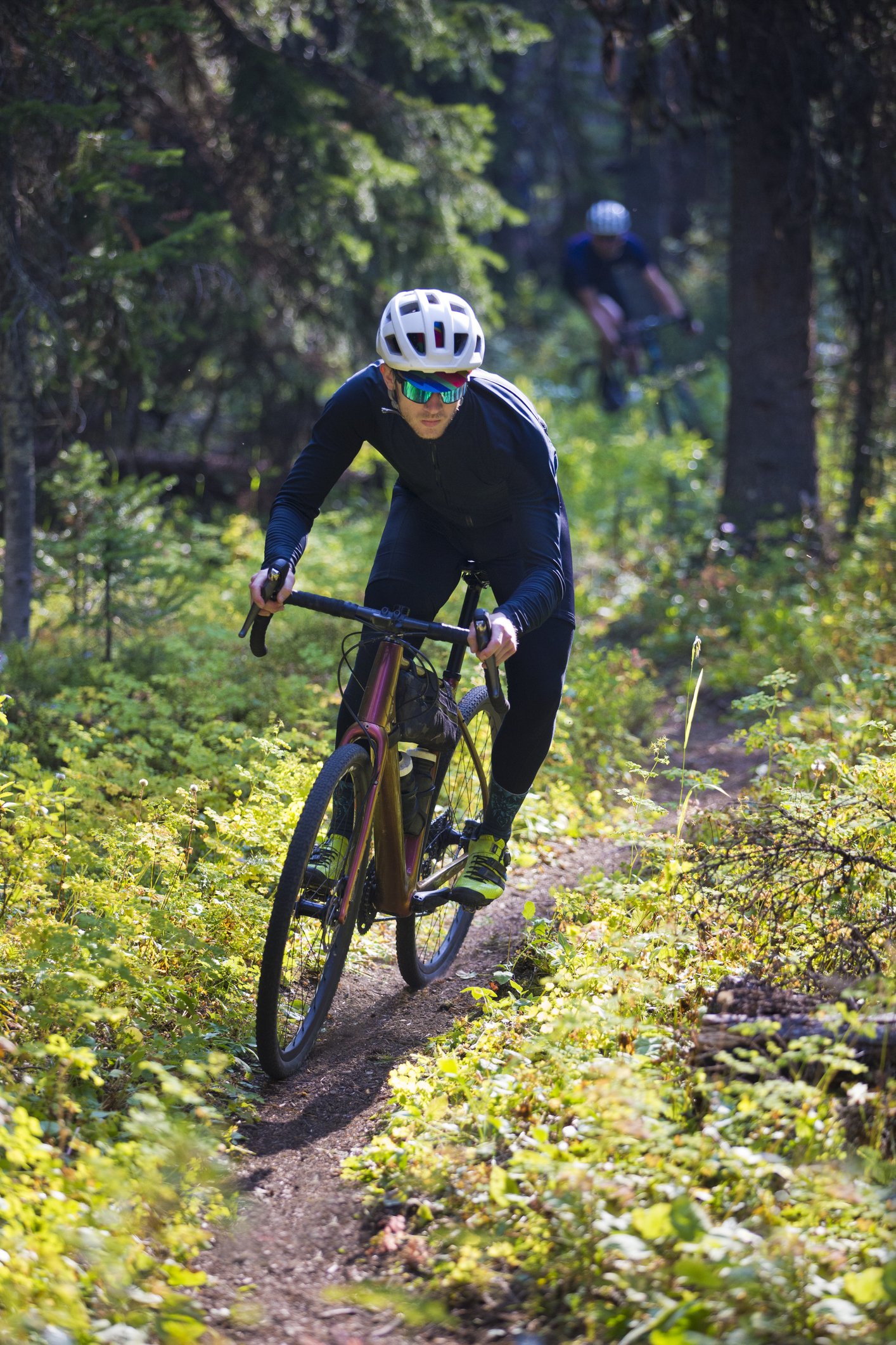 A man rides his gravel bike on a singletrack trail in British Columbia, Canada. He is followed by another rider in the background. Gravel bikes are similar to cyclo-cross bicycles with sturdy wheels and tires suited to riding on rough terrain. He has a frame bag attached to his bike to carry extra gear.