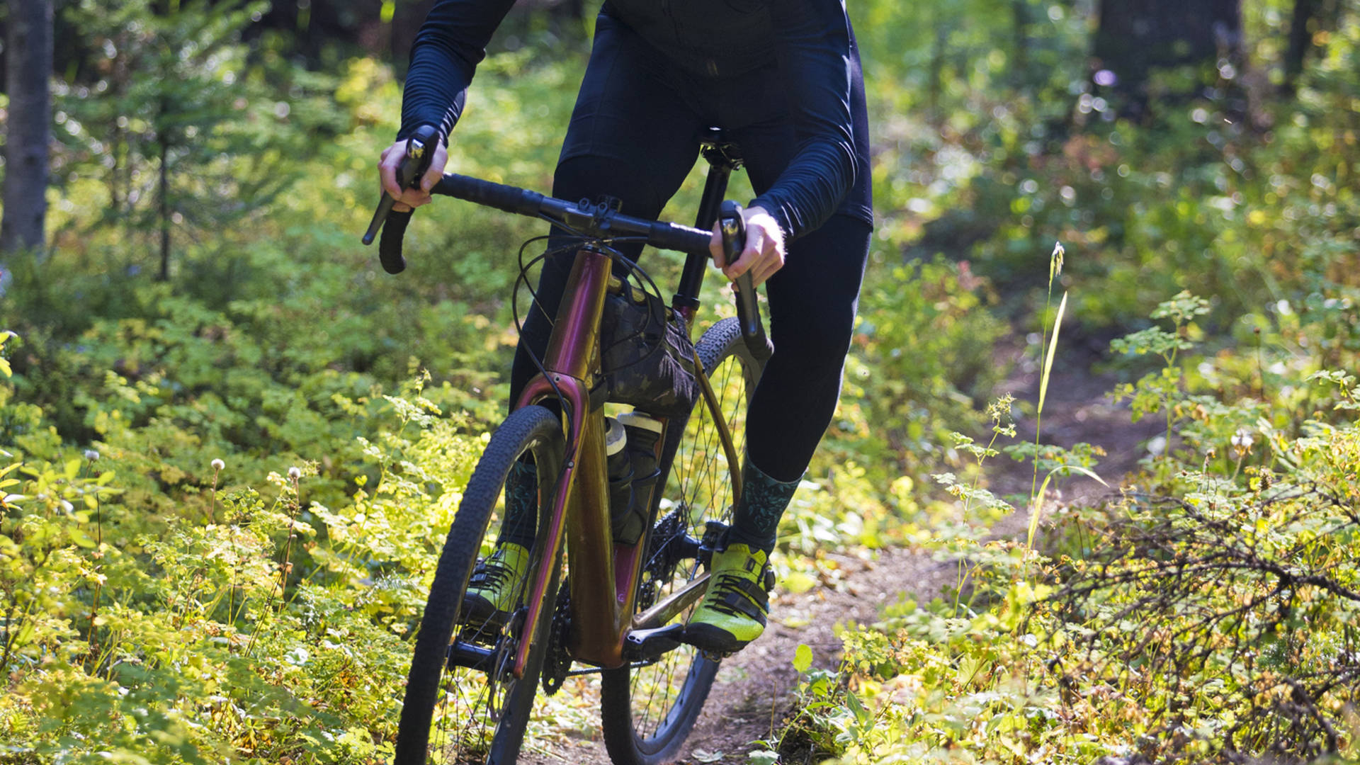 A man rides his gravel bike on a singletrack trail in British Columbia, Canada. He is followed by another rider in the background. Gravel bikes are similar to cyclo-cross bicycles with sturdy wheels and tires suited to riding on rough terrain. He has a frame bag attached to his bike to carry extra gear.