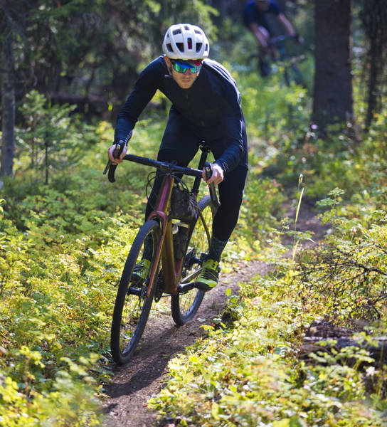 A man rides his gravel bike on a singletrack trail in British Columbia, Canada. He is followed by another rider in the background. Gravel bikes are similar to cyclo-cross bicycles with sturdy wheels and tires suited to riding on rough terrain. He has a frame bag attached to his bike to carry extra gear.