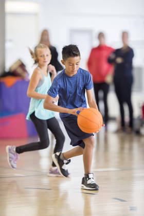 A multi-ethnic group of elementary age children are playing basketball together in the gym. They are dribbling the ball up the court.