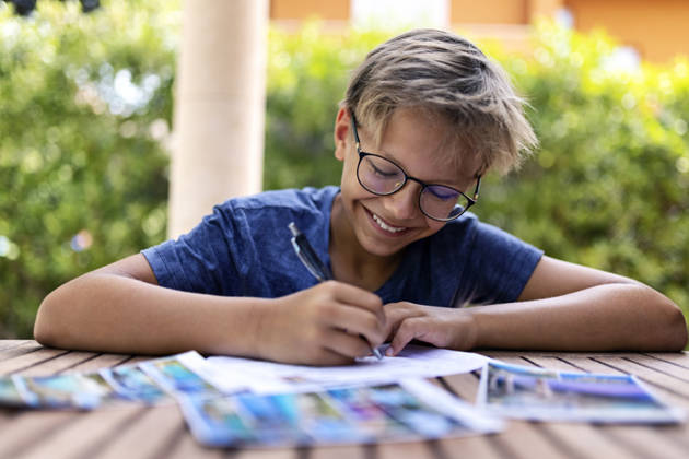 Teenager boy writing postcards to friends and family. He is sitting by a table in the backyard. Toothy smile, one person, laughing..Canon R5