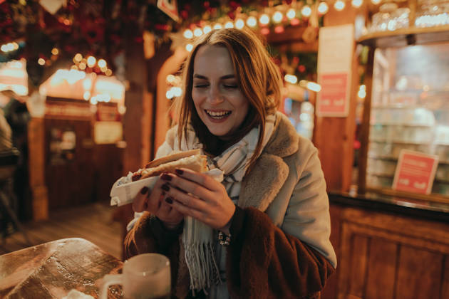 Happy young woman enjoying a Christmas market outdoors in the city and eating a sausage hot dog on a lovely winter night