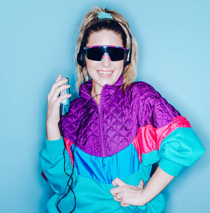A woman wearing clothing styled after the 1980's and 1990's listens to music on her personal cassette tape player in front of a large bright blue background. Shot with a ring flash.