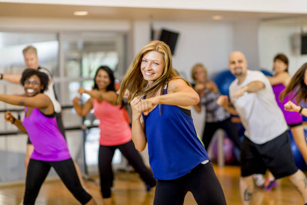 A woman is leading a multi-ethnic boxing class, everyone is wearing athletic clothing and is throwing a punch together - they are smiling and looking at the camera.