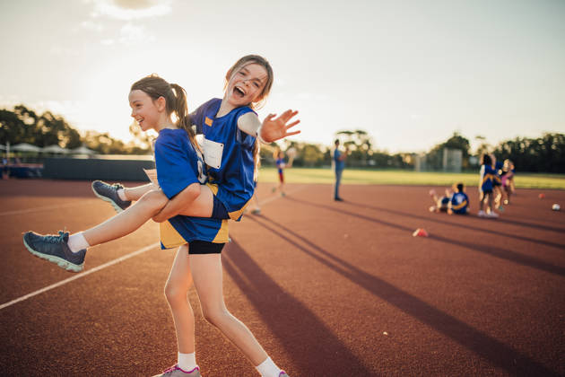 Two girls are messing around at athletics club on the running track. One is giving the other a piggy back and she is reaching out for the camera.