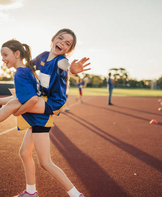 Two girls are messing around at athletics club on the running track. One is giving the other a piggy back and she is reaching out for the camera.
