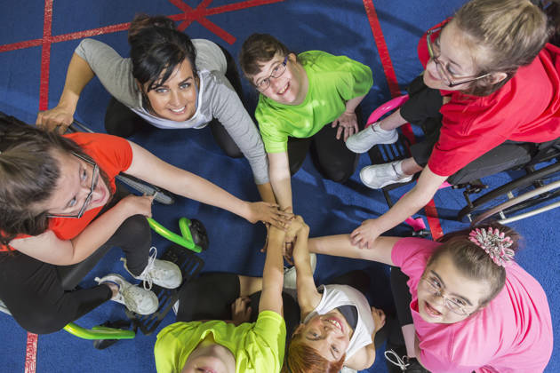 Overhead view of a group of special needs teenagers and young women with instructor, showing team spirit. They are sitting in a circle on a gym floor, hands in the center. Several of the girls have downs syndrome and two are in wheelchairs.