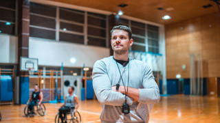 Portrait of a wheelchair basketball coach standing with his arms crossed and looking at camera while his team is practicing in the background.