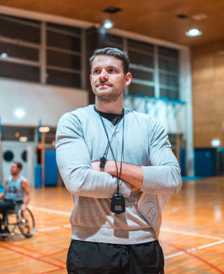 Portrait of a wheelchair basketball coach standing with his arms crossed and looking at camera while his team is practicing in the background.
