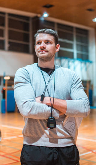 Portrait of a wheelchair basketball coach standing with his arms crossed and looking at camera while his team is practicing in the background.