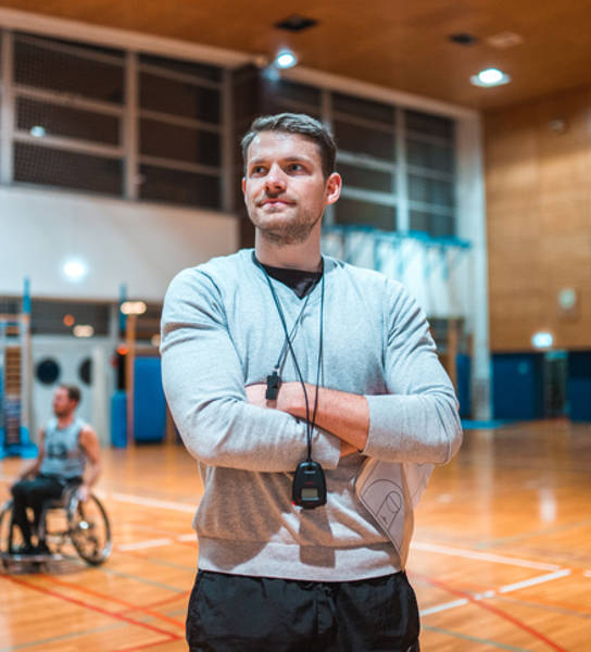 Portrait of a wheelchair basketball coach standing with his arms crossed and looking at camera while his team is practicing in the background.