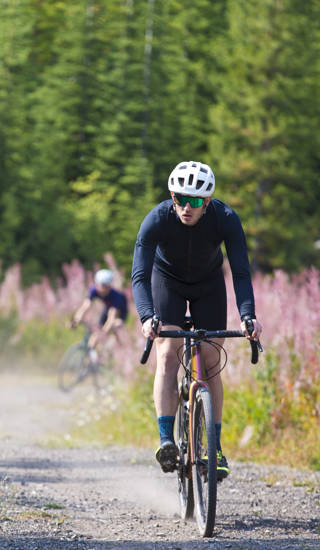 A man goes for a ride on his gravel bicycle in the Rocky Mountains of Canada. He is followed by two other riders in the background. Gravel bikes are similar to cyclo-cross bicycles with sturdy wheels and tires for riding on rough terrain. His bike has a large frame bag to carry extra gear.