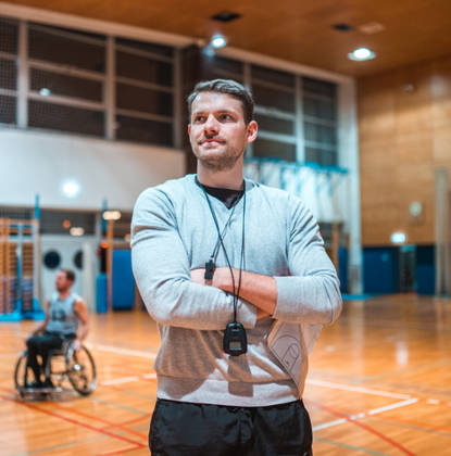 Portrait of a wheelchair basketball coach standing with his arms crossed and looking at camera while his team is practicing in the background.